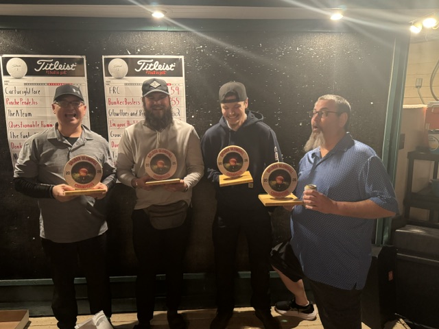 Four winners posing with trophies by the golf scoreboard