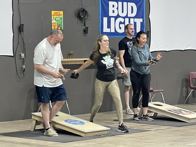Player mid-toss with laughing teammates at cornhole boards